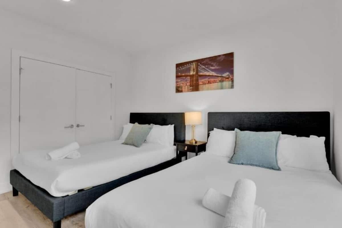 A hotel room with two beds, a nightstand with a lamp, a painting of a bridge, and a white door.