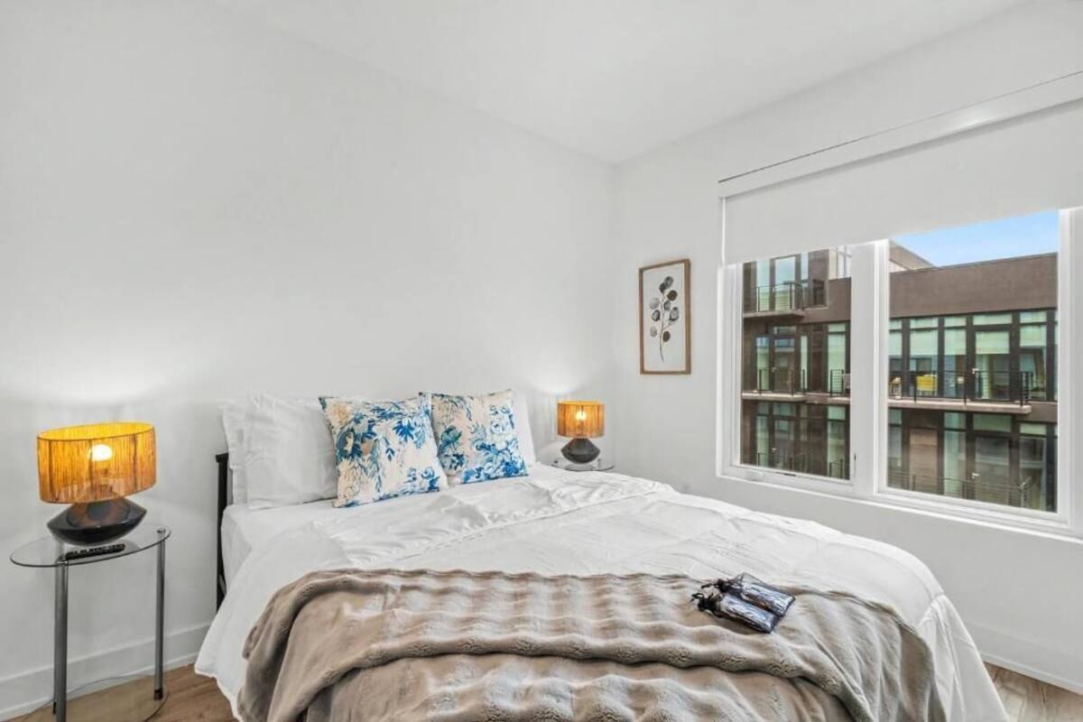 A bedroom with a bed, two bedside lamps, a window with a view of buildings, and a framed picture on the wall.