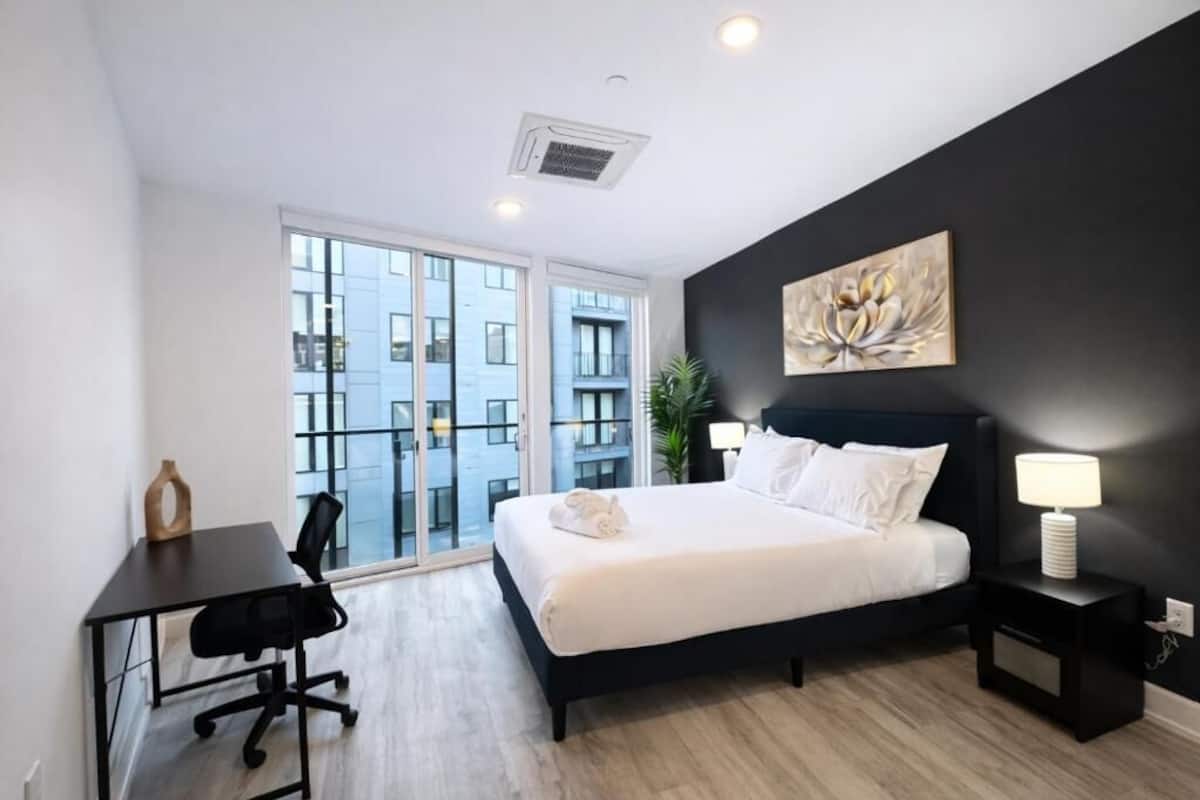 A modern hotel room with a large bed, a desk, a chair, a nightstand, a lamp, and a view of the city through large windows.