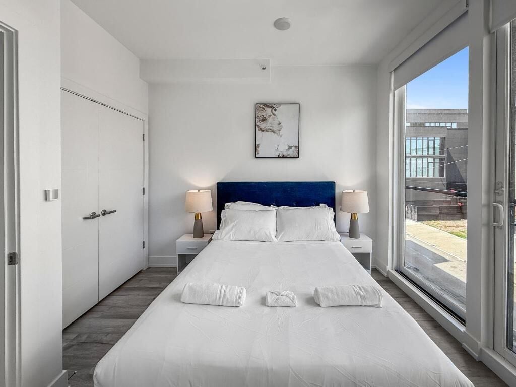 Ett modernt hotellrum med en stor säng, vita sängkläder, en blå sänggavel, sänglampor och utsikt genom en glasdörr.