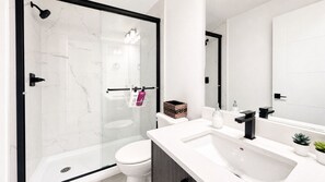 Un baño moderno con cabina de ducha de vidrio, inodoro blanco, lavabo blanco con grifo negro y espejo sobre el lavabo.