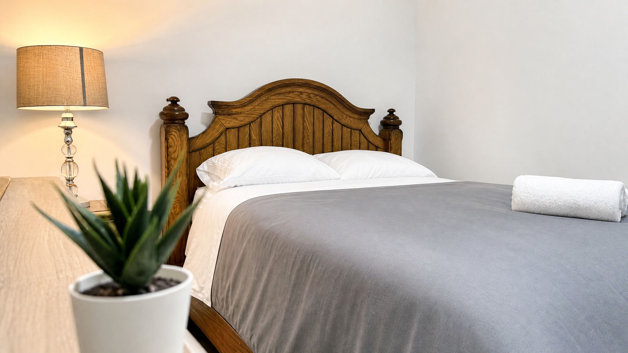 Un lit bien fait, avec une tête de lit en bois, une lampe de chevet et une plante en pot.