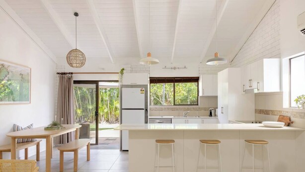 A modern kitchen with white cabinetry, a central island with stools, a dining area with a table and benches, and a view of greenery outside.