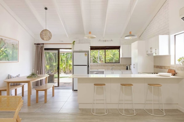 A modern kitchen with white cabinetry, a central island with stools, a dining area with a table and benches, and a view of greenery outside.