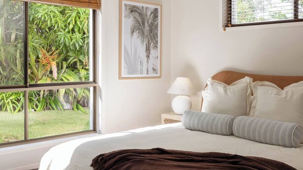 Ein Schlafzimmer mit großem Fenster, einem Bett mit weißer Bettwäsche, einer braunen Decke und einem gerahmten Bild an der Wand.
