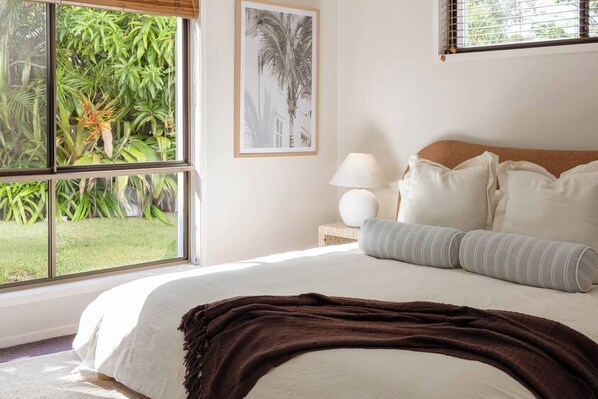 Ein Schlafzimmer mit großem Fenster, einem Bett mit weißer Bettwäsche, einer braunen Decke und einem gerahmten Bild an der Wand.