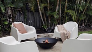 Eine Terrasse mit weißen Stühlen, einer schwarzen Feuerstelle in Schalenform und üppigen tropischen Pflanzen.