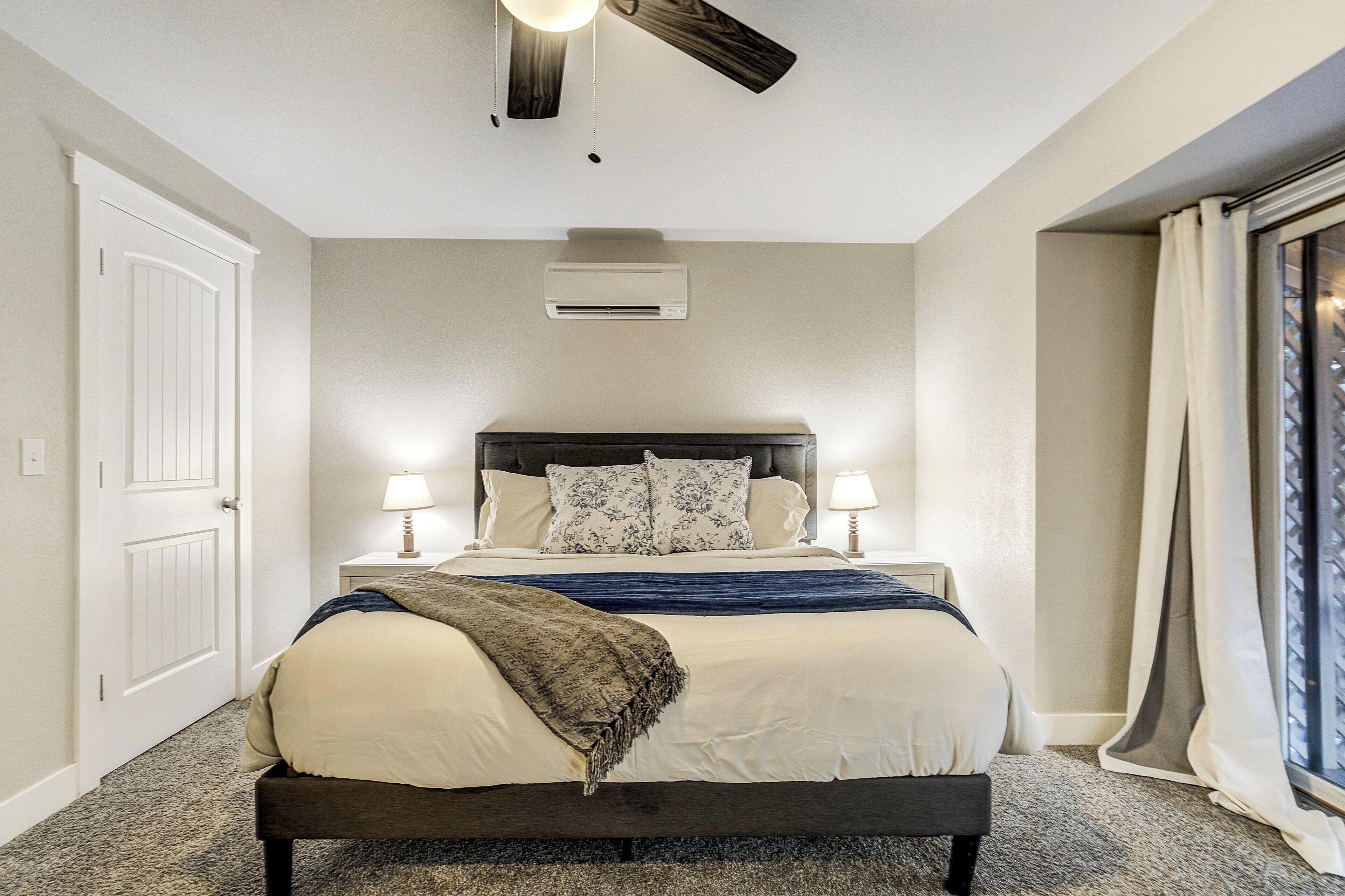 Ein Schlafzimmer mit einem Bett, Nachttischlampen, einem Deckenventilator, einer Klimaanlage und einem Fenster mit Vorhängen.