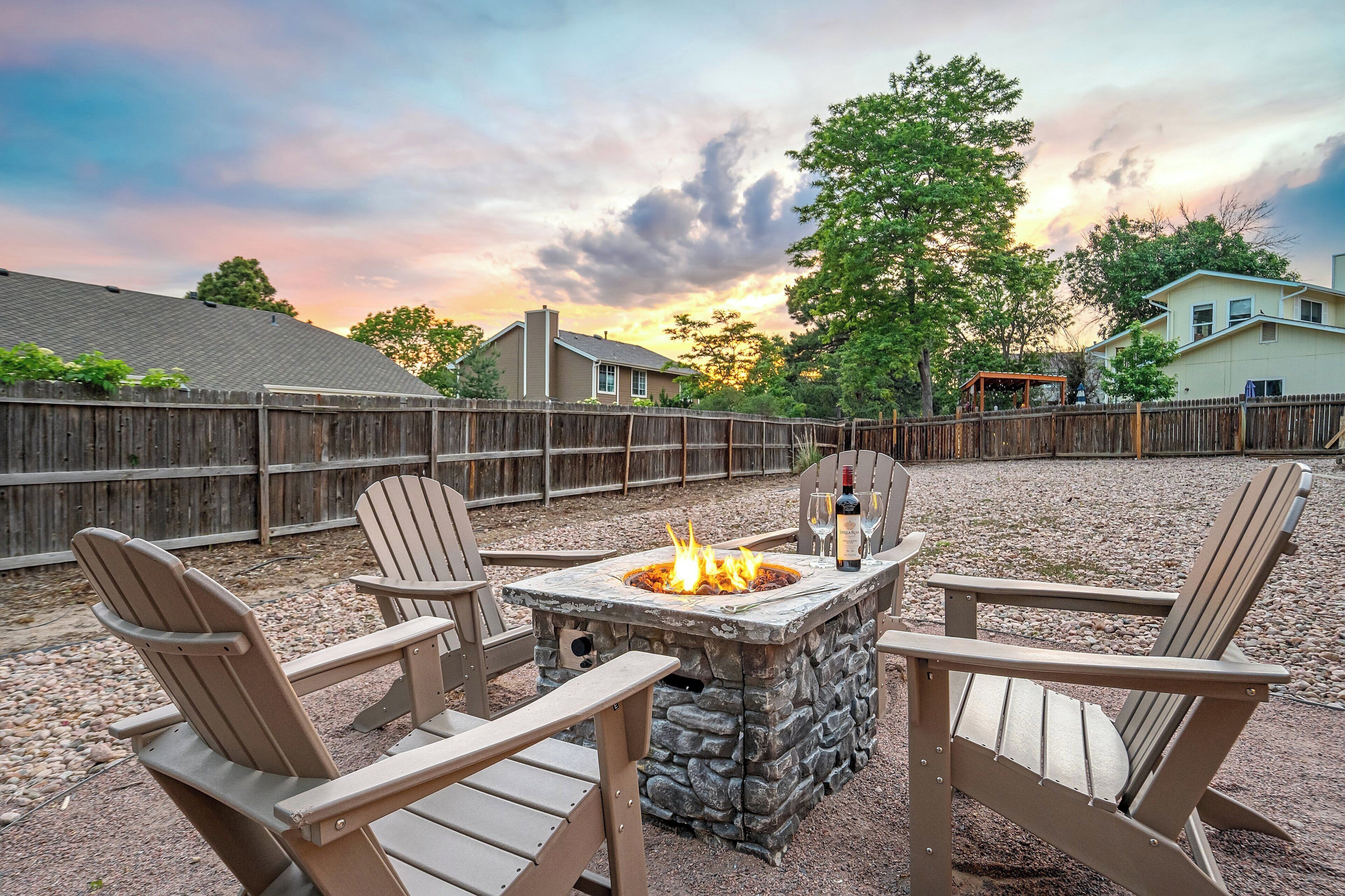 A stone fire pit with chairs around it, set in a backyard with a wooden fence and houses in the background.