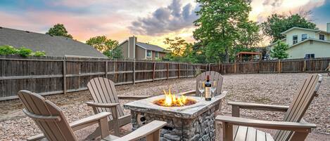 A stone fire pit with chairs around it, set in a backyard with a wooden fence and houses in the background.