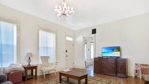 Ein Wohnzimmer mit einer Kronleuchter, einem Fernseher, einem Sofa, einem Sessel, einem Tisch und einem gemusterten Teppich.