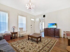Ein Wohnzimmer mit einer Kronleuchter, einem Fernseher, einem Sofa, einem Sessel, einem Tisch und einem gemusterten Teppich.