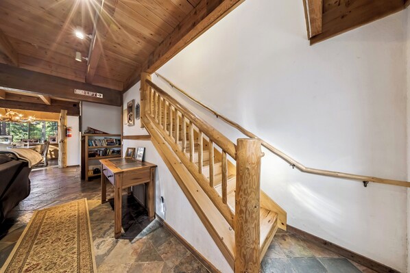 Eine Holztreppe mit Geländer, ein Schild mit der Aufschrift 'Skilift', ein Holztisch, eine Couch und ein Teppich in einem Raum mit Steinböden und Holzbalken.