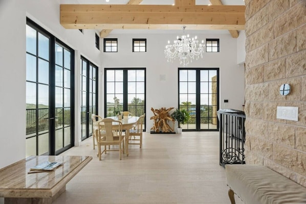 Una habitación amplia con ventanas grandes, techo de madera, una araña y una zona de comedor con una mesa y sillas.