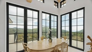 Un espace repas comprenant une grande table et des chaises, de grandes fenêtres offrant une vue sur les montagnes, et une sculpture en bois posée sur le sol.