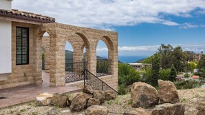 Une construction en pierre avec des arcades, un balcon doté d’une balustrade en métal, et une vue sur la mer et les collines.