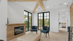 Un salon spacieux doté d’une cheminée en pierre, de deux fauteuils bleu foncé, d’une table basse en bois et de grandes fenêtres offrant une vue sur l’extérieur.