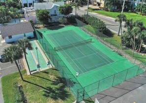 Ein Tennisplatz, von einem Zaun umgeben, mit einem Haus und Palmen im Hintergrund.