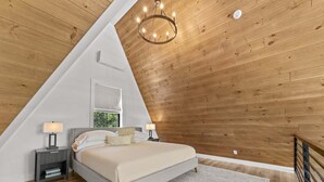 A bedroom with a sloped ceiling, a bed, bedside lamps, a window, and a chandelier. - Fernwood: modern cabin with hot tub, putting green, pickleball (Hammondsport)