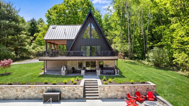 A modern two-story house with a balcony, stone steps leading to the entrance, and outdoor seating. - Fernwood: modern cabin with hot tub, putting green, pickleball (Hammondsport)