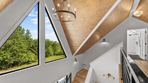 A modern interior with a wooden ceiling, large windows, a staircase, and a view of greenery outside. - Fernwood: modern cabin with hot tub, putting green, pickleball (Hammondsport)