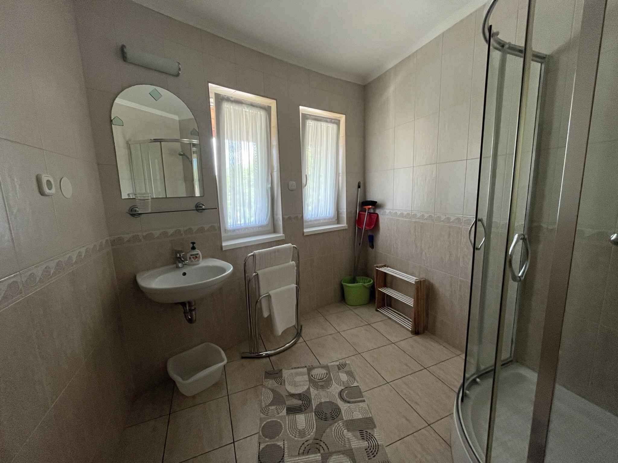 Une salle de bain avec un lavabo, un miroir, deux fenêtres avec des rideaux, une douche et un carrelage décoratif.