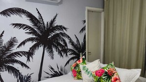 Habitació d'hotel amb un llit, un mural tropical, un coixí floral, una tovallola blanca i un aparell d'aire condicionat.
