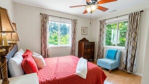 Un dormitorio con cama, una silla, ventilador de techo y dos ventanas con cortinas.