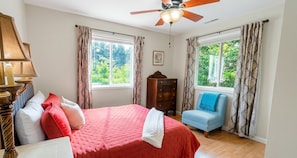 A bedroom with a bed, a chair, a ceiling fan, and two windows with curtains.