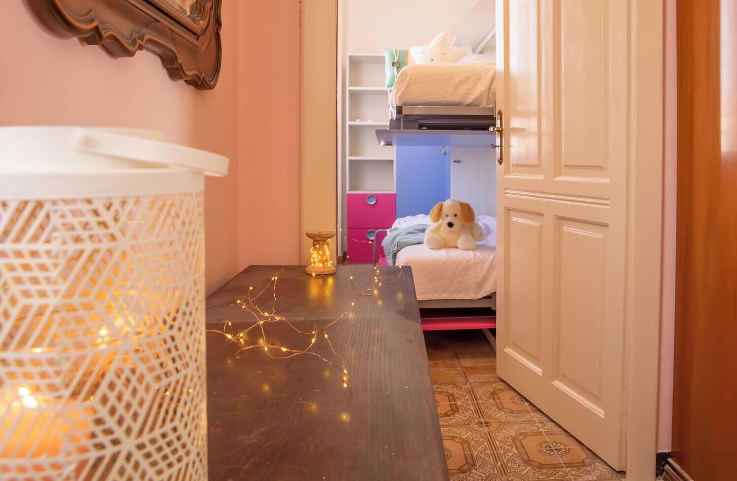 Een kamer met een stapelbed, een klein tafeltje met lichtjes, een decoratief wandobject en een deur.