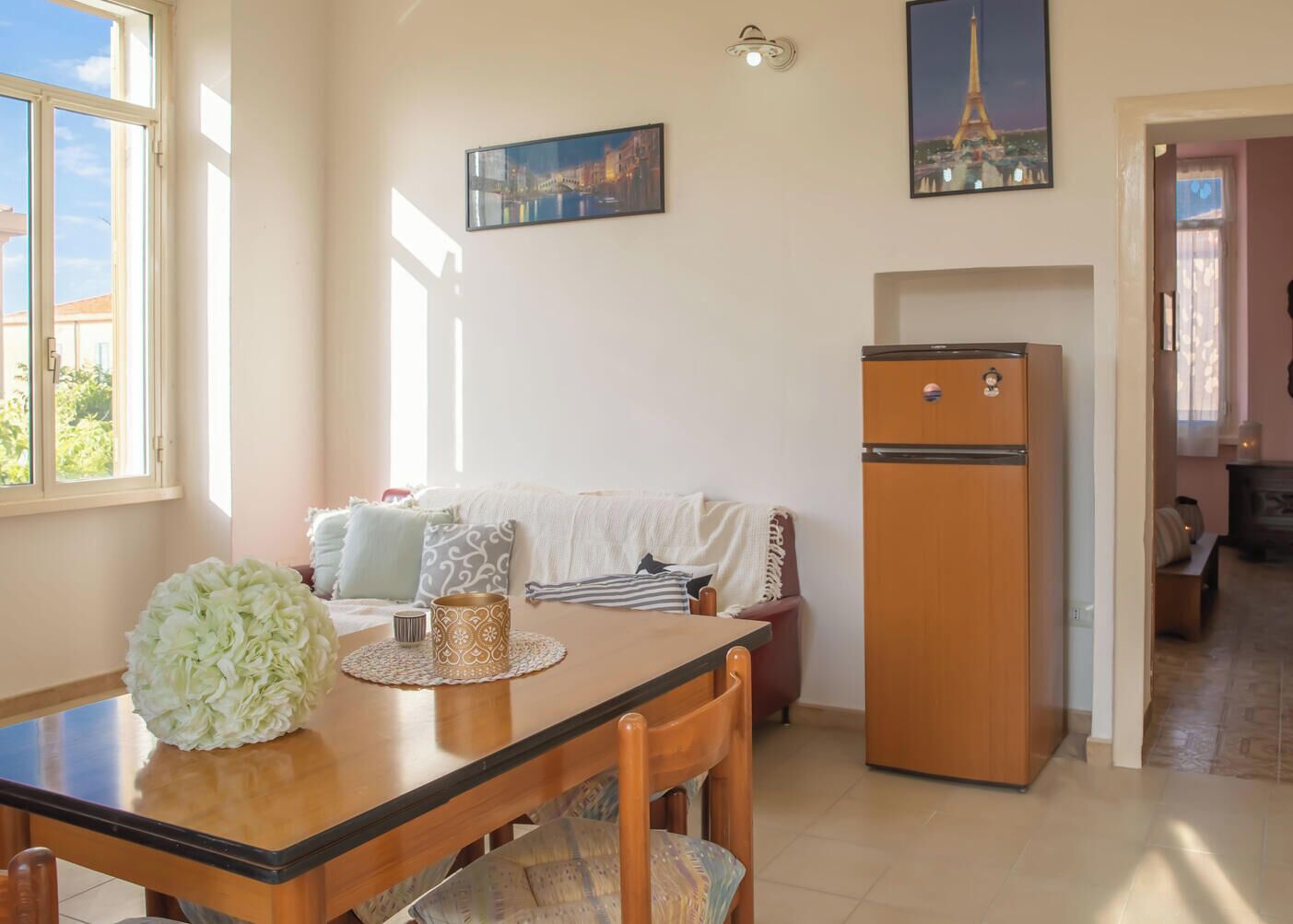 A cozy living space with a dining table, a sofa, a refrigerator, and a window with a view of greenery.