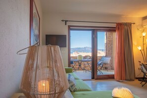 A room with a green sofa, a table with chairs, a TV on the wall, and a view through the sliding glass door. - The Port of Dreams - Porto Rotondo (Porto Rotondo)