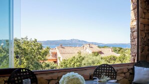 Uma mesa com um copo d'água, um arranjo de flores brancas e um castiçal, com uma parede de pedra ao fundo e vista para edifícios e árvores.