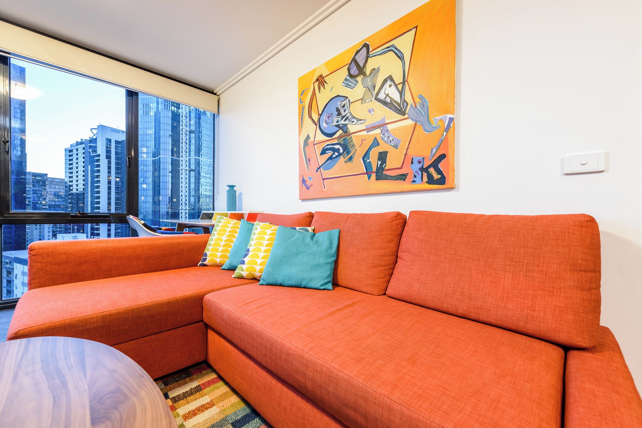 A modern living room with a large orange sectional sofa, colorful pillows, a city view through large windows, and a vibrant abstract painting on the wall.