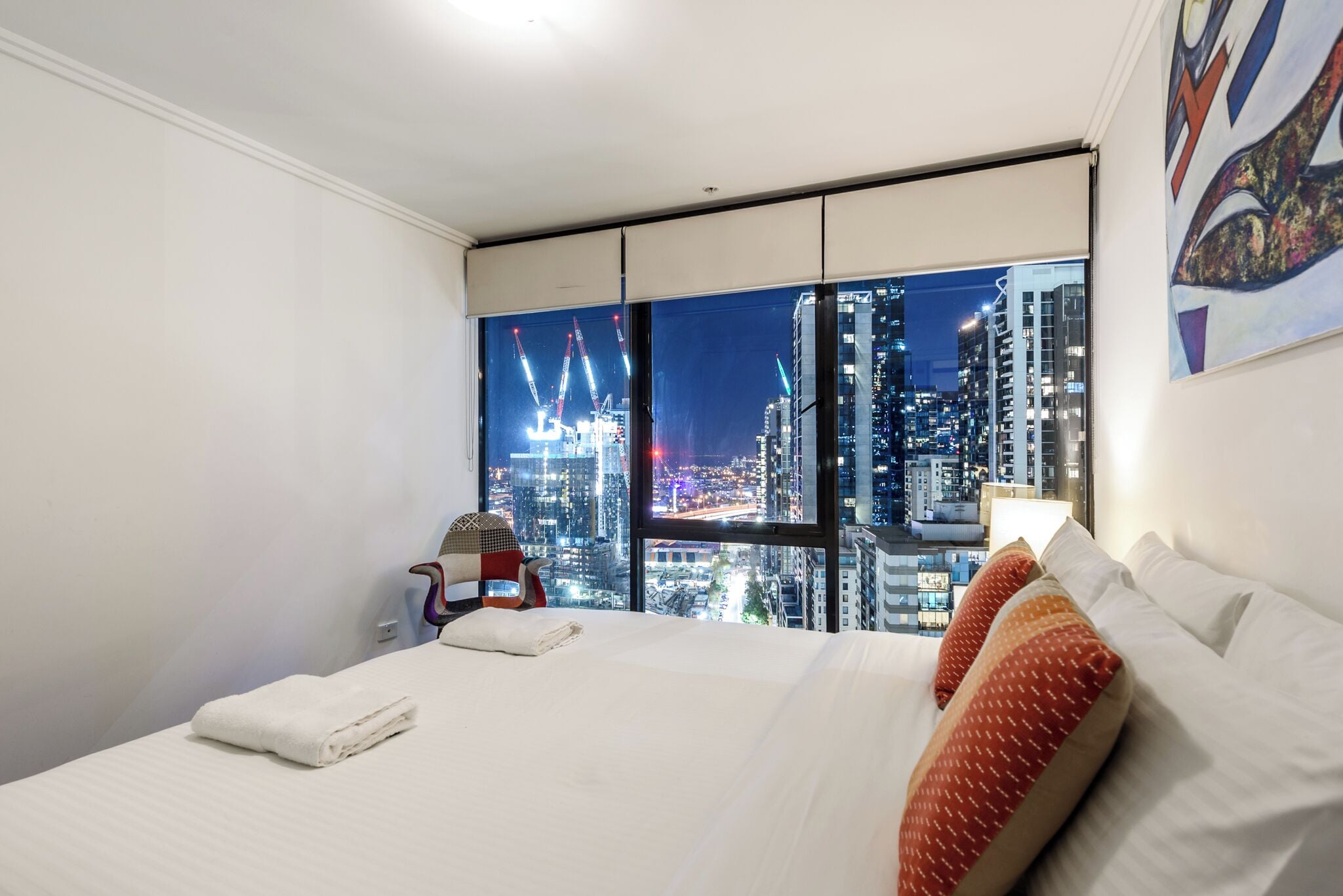 Quarto de hotel com uma janela ampla que oferece vista para a cidade, uma cama bem arrumada e uma luminária fixada na parede.