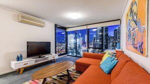 Un salón moderno con un televisor grande, un sofá naranja y vistas a la ciudad a través de amplias ventanas.
