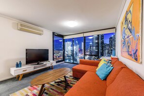 A modern living room with a large TV, orange sofa, and a city view through large windows. - Exclusive Stays - Southpoint (Southbank)