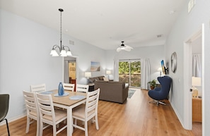 Ein geräumiges Wohnzimmer mit Esstisch, Stühlen, einer Couch und einem blauen Sessel. Es gibt Deckenventilatoren, einen Balkon mit Blick und eine Tür zu einem weiteren Raum.