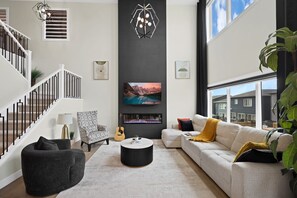 A modern living room with a staircase, a fireplace, a sofa, and a large window. - Designer-Furnished Former Show Home | AC | BBQ (Calgary)