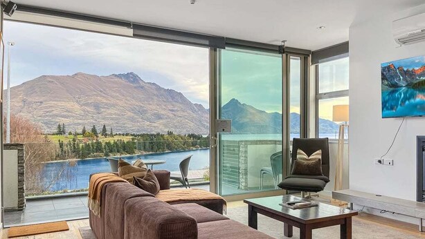 Ein modernes Wohnzimmer mit einem großen Glasfenster, das auf einen Balkon mit Blick auf einen See und Berge führt.