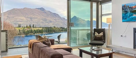 A modern living room with a large glass door leading to a balcony overlooking a lake and mountains.