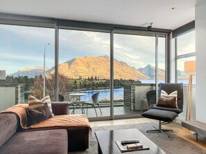 En moderne stue med et stort skydedørsvindue, der fører ud til en balkon med udsigt over bjerge og en sø.