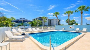 Una piscina rectangular con sillas de descanso blancas y una cerca blanca, rodeada de palmeras y un edificio con balcones.