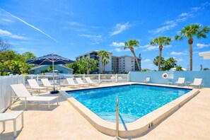 A rectangular pool with white lounge chairs and a white fence, surrounded by palm trees and a building with balconies. - Lido Key 2 bed Heated Pool and al fresco shower (Sarasota)
