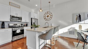 A modern kitchen with white cabinets, stainless steel appliances, a central island with bar stools, a wall clock, and a dining area with a patterned rug. - Apartmint at Germantown Ave Philadelphia (Philadelphia)