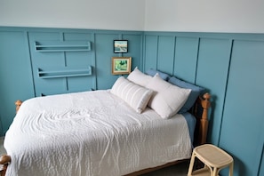 A bed with a white quilt, a wooden headboard, and a small wicker chair. There are two framed pictures on the wall. - Rooftop Bay Views - Downtown Lakehouse Sleeps 8 (Sandusky)