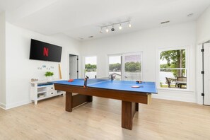 Une pièce avec une table de ping-pong, un téléviseur fixé au mur et de grandes fenêtres donnant sur un plan d’eau.