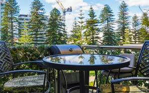 A patio with a round table and chairs, surrounded by greenery and tall trees.