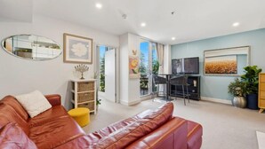 A living room with a brown leather sofa, a small yellow ottoman, a fireplace, a television, and a large window. - Park View Hideaway (Glenelg)