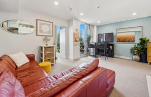 A living room with a brown leather sofa, a small yellow ottoman, a fireplace, a television, and a large window. - Park View Hideaway (Glenelg)